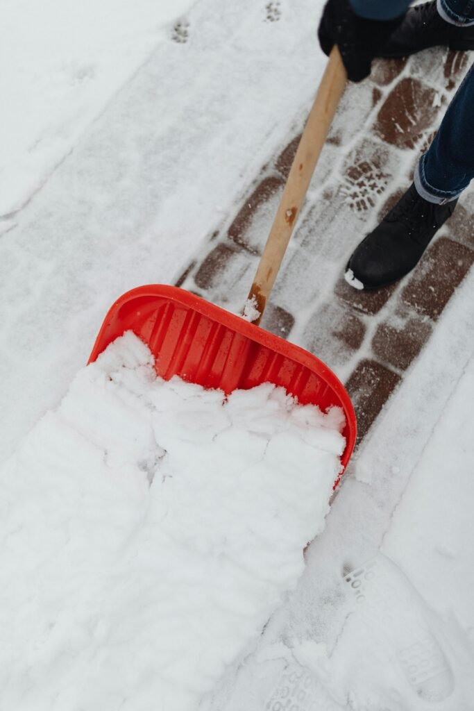 A person uses a red shovel to clear snow from a cobblestone sidewalk during winter.